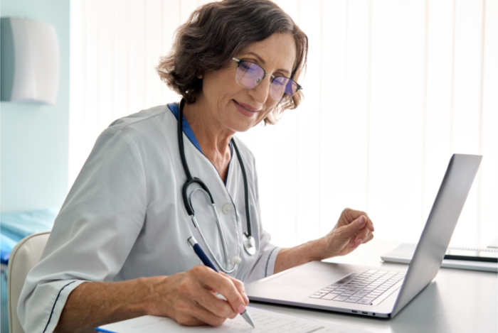 Woman doctor reviewing patient notes on a laptop writing on a clipboard