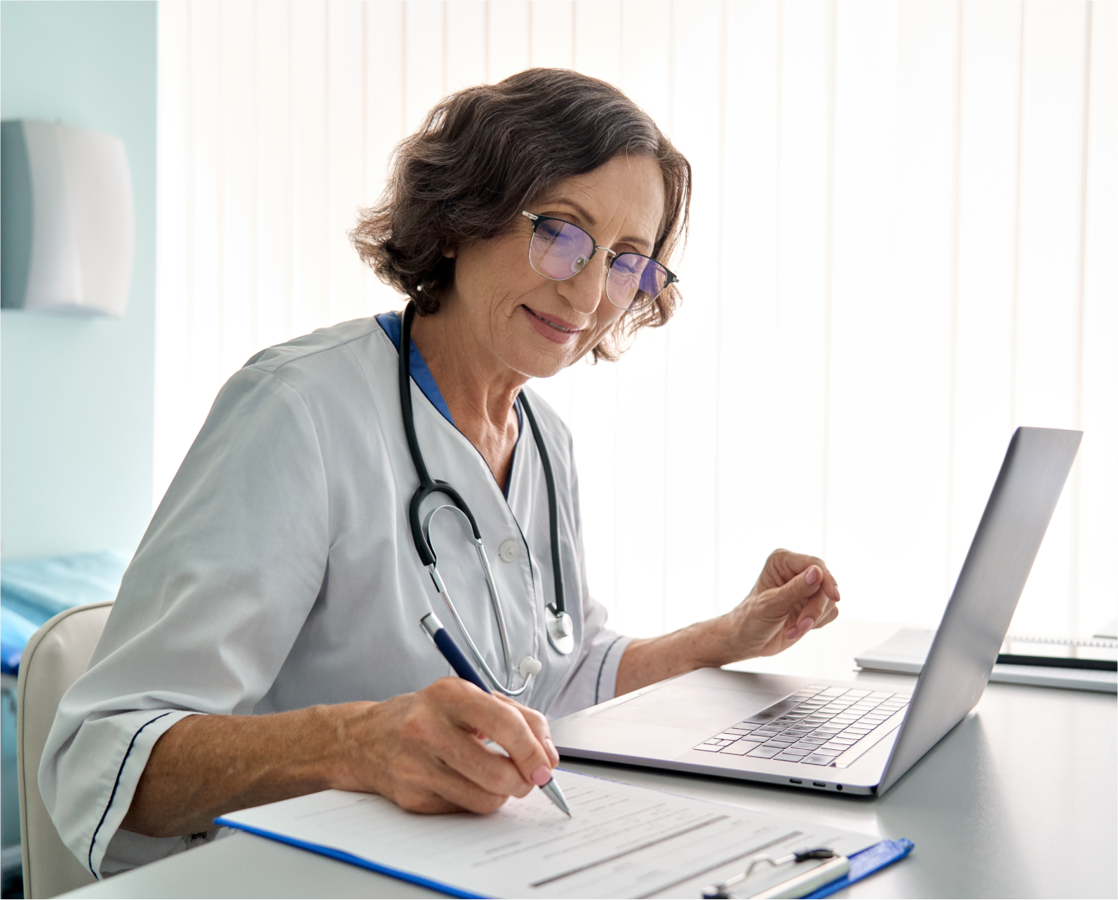 Woman doctor reviewing patient notes on a laptop writing on a clipboard