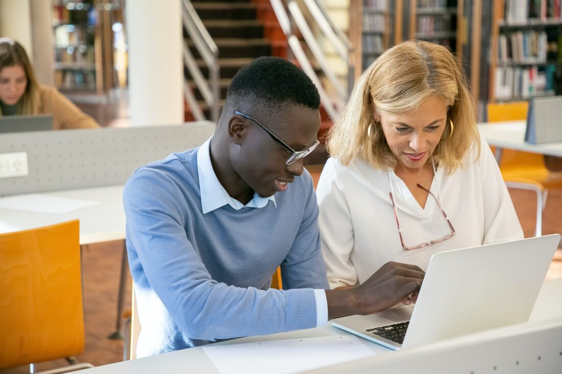 Student working with teacher in a library on a laptop together